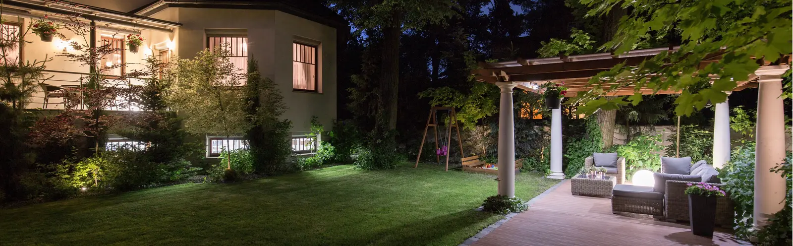 Hage og terrasse om kvelden med små lyspunkter langs bed, plen og pergola