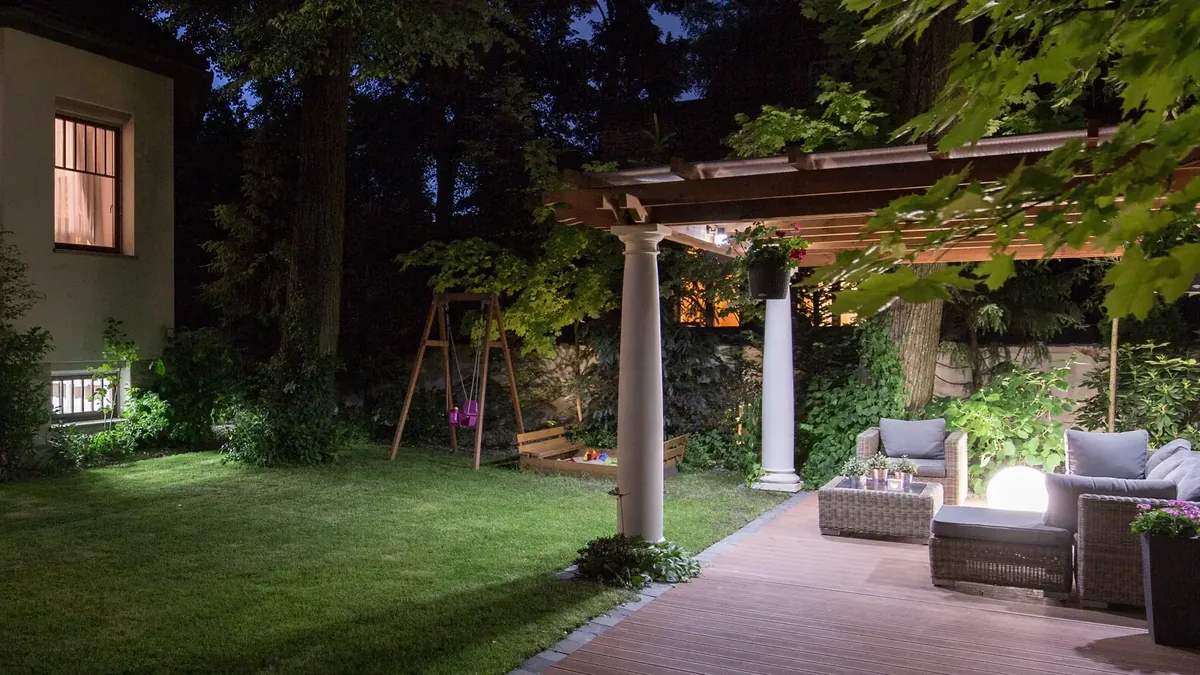 Terrasse med pergola, utemøbler og lyskule om kvelden, med lys i bedet langs plenen