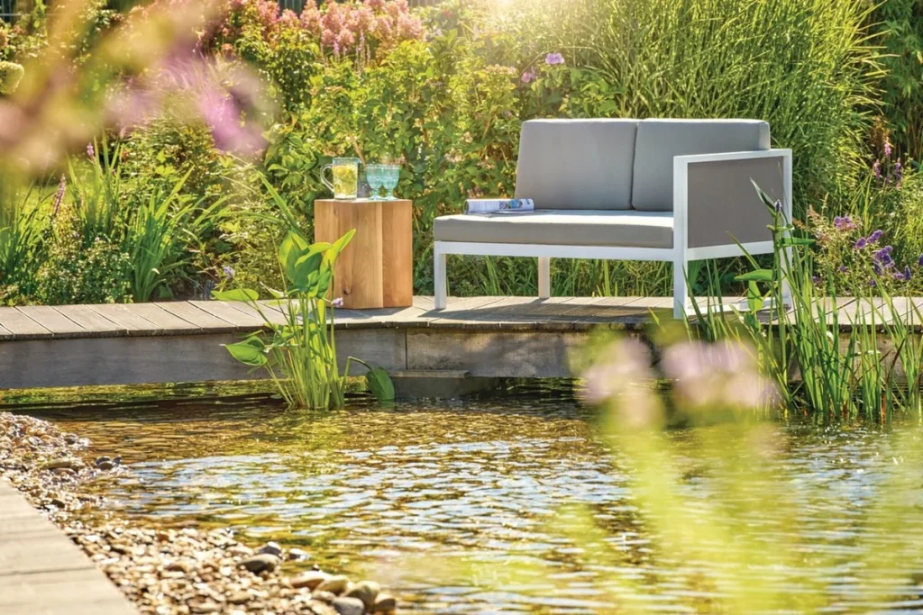 Hagedam med klart vann foran en terrasse med sofa og blomstrende stauder