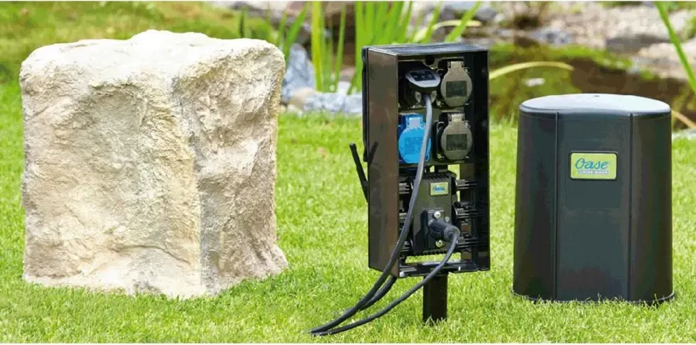 El-skapet OASE FM Master WLAN EGC på plenen ved dammen, med steinlokk ved siden av