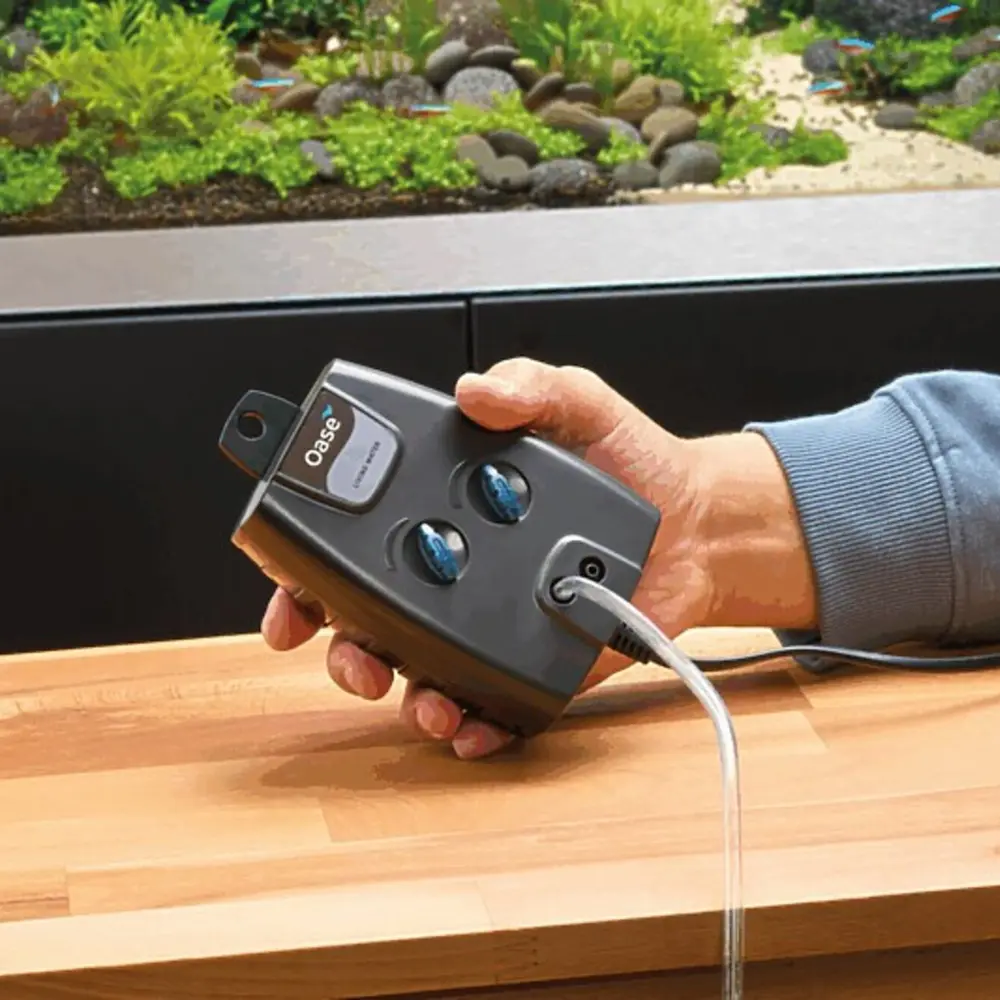 OASE OxyMax luftpumpe med to uttak og to ventiler holdes i hånden foran et akvarium